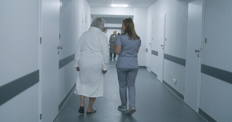 Female doctor, nurse with digital tablet walks along the clinic corridor with elderly woman, helps patient to get to hospital ward after procedures. Medical staff, patients in medical center hallway.