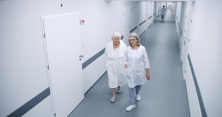 Bright clinic corridor: Female doctor and elderly woman walk down the hallway to hospital ward,...