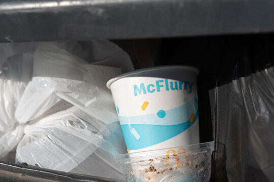 discarded McFlurry cup and plastic dome in a recycling bin with bags of garbage in strong sunlight