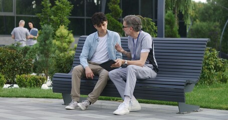 Mature doctor with tablet in hands sits with middle aged patient on bench and consults him....