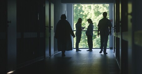 Fototapeta premium Hospital hallway: Doctors and professional medics walk. Nurse with digital tablet stands with adult woman near window and talks. Medical staff and patients in dark clinic or medical center corridor.