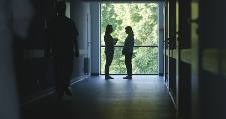 Clinic corridor: Doctors and professional medics walk. Nurse with digital tablet comes to female patient standing near window. Medical staff and patients in dark hospital or medical center hallway.