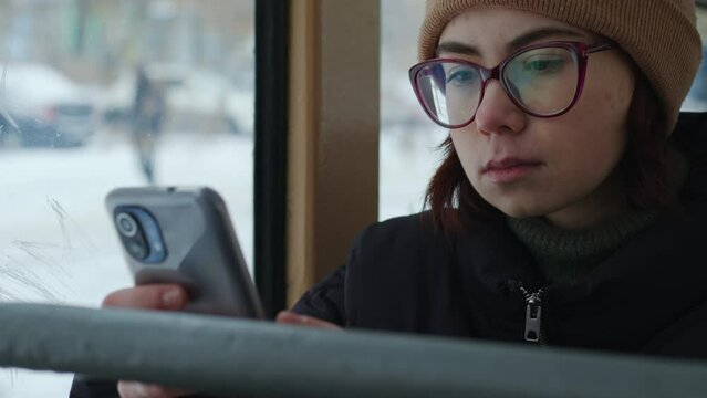 Sad girl in puffer jacket sits at bus, looks to cellphone. Young woman chatting with friend on social networks while riding in tram. Student typing on smartphone in public transport. Side view