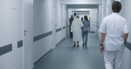 Female doctor, nurse with digital tablet walks along the clinic corridor with elderly woman, helps patient to get to hospital ward after procedures. Medical staff, patients in medical center hallway.