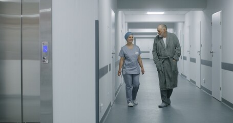 Nurse in uniform walks hospital corridor with aged man and comes to lift door. Female healthcare specialist meets elderly patient in medical center hallway. Medical personnel work in modern clinic.