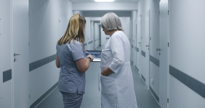 Nurse shows MRI or CT brain scan image to female doctor using digital tablet. Medics stand and walk along the modern clinic corridor, discuss work. Medical staff in hospital or medical center hallway.