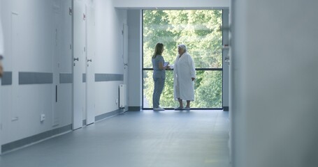 Hospital hallway: Doctors and professional medics walk. Nurse with digital tablet comes to elderly female patient standing near window. Medical staff and patients in clinic or medical center corridor.