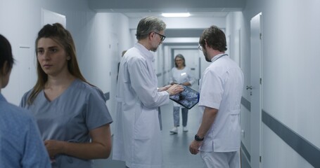 Mature doctor shows MRI, CT brain scan image to colleague using digital tablet. Professional medics stand and walk along the clinic corridor, discuss work. Medical staff, patients in hospital hallway.