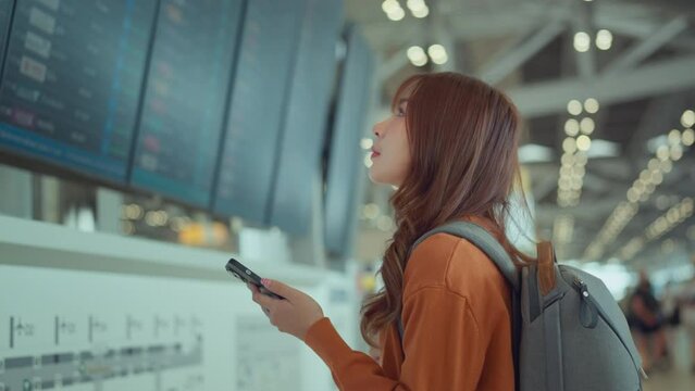 Happy asian woman traveller checking flight schedule departures board in airport terminal hall in front of check in counters. Tourist journey trip concept