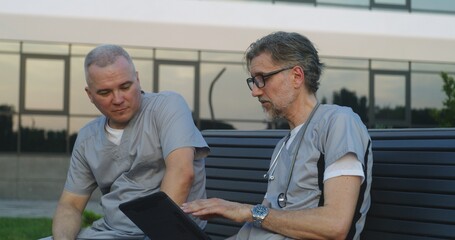 Professional doctors in uniform sit on bench near modern hospital. Medics discuss work, watch medical tests results on digital tablet. Medical staff of clinic or medical center. Healthcare system.