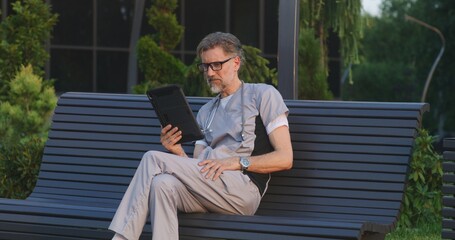 Fototapeta premium Doctor in uniform sits on bench outside modern hospital, talks to patient about treatment during online appointment using digital tablet. Medical staff of clinic or medical center. Healthcare system.