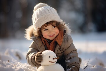 Winterspaß mit Kindern; kleiner Junge trägt braune Handschuhe und dicke Winterjacke und weiße Mütze, baut einen Schneemann im Schnee und lächelt  - obrazy, fototapety, plakaty