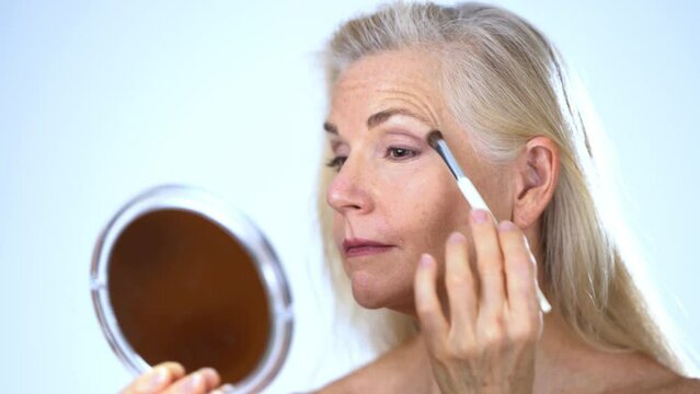 Closeup Of Mature Middle-aged Elderly Woman Putting On Eyeliner, Concealer, Eye Shadow Hygienic Makeup In Mirror Apply Facial Powder, Face Cosmetics Isolated On White Studio Background