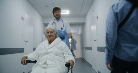 Young doctor in glasses carries senior patient in wheelchair down hospital corridor. Old woman talks to medic and smiles. Mature doctor asks female patient about her health. Medical staff at work.