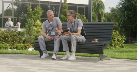 Two doctors sit on bench and discuss patient treatment. Health care specialists look at client tests results using tablet. Mature man helps colleague with making diagnosis. Medical staff work outdoor.