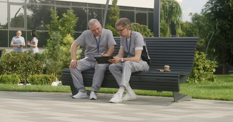 Two doctors sit on bench and discuss patient treatment. Health care specialists look at client tests results using tablet. Mature man helps colleague with making diagnosis. Medical staff work outdoor.