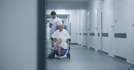 Male doctor stands with elderly woman in wheelchair in clinic corridor, uses mobile phone. Medic with female patient near operating or procedures room. Medical staff and patients in hospital hallway.