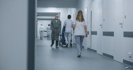 Fototapeta premium Mature doctor stands in clinic corridor with nurse and patient. Professional medic talks with female colleague and elderly woman, uses digital tablet. Medical staff and patients in hospital hallway.