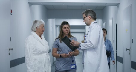 Fototapeta premium Mature doctor stands in clinic corridor with nurse and patient. Professional medic talks with female colleague and elderly woman, uses digital tablet. Medical staff and patients in hospital hallway.