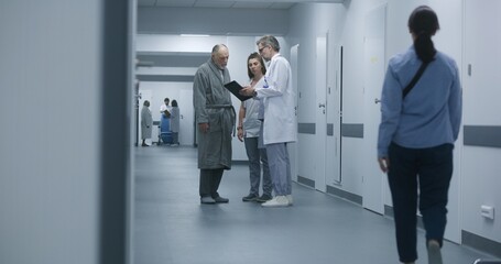 Mature doctor stands in clinic corridor with nurse and patient. Professional medic talks with female colleague and elderly woman, uses digital tablet. Medical staff and patients in hospital hallway.
