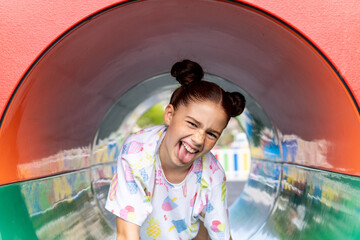Portrait of a crazy red haired girl with freckles laughing and sticking her tongue out inside a tube in the playground, concept of happiness and joy, carrying a backpack.