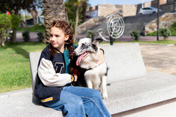 Red haired girl with  freckles smiling, wearing a coloured jacket, sitting in the park with her black and white dog