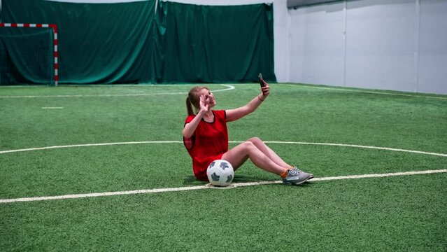 smiling young soccer girl blogger taking photo on phone taking selfie with ball on soccer field