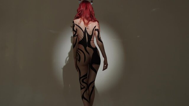 A devil girl with horns on her head in a sexy tight costume dances beautifully in the studio on a gray background.