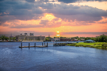 A beautiful summer landscape along Cape Fear River with flowing water, lush green trees and grass, a pergola with people, boats and buildings along the banks of the river in Wilmington North Carolina