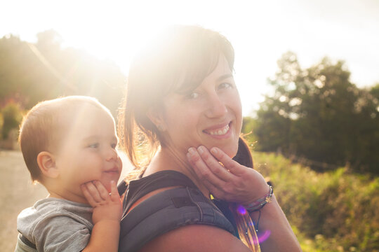 Close-up of a mother with her child carried in a backpack in a backlit sunset.