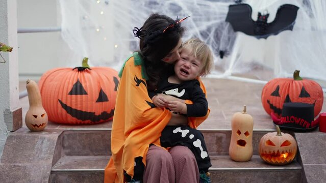 a child dressed as a skeleton for Halloween is crying from fright and a mother dressed as a pumpkin calms him down on the porch of the house against the background of Halloween props, bats, pumpkins