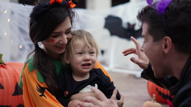 a child dressed as a skeleton for Halloween is crying from fright and a mother dressed as a pumpkin calms him down on the porch of the house against the background of Halloween props, bats, pumpkins