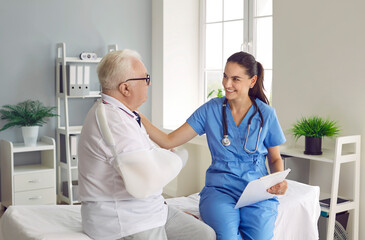 Smiling doctor supporting elder patient with arm injury. Young woman in blue uniform scrubs together with old man with broken arm in sling sitting on medical bed and talking. Injury treatment concept