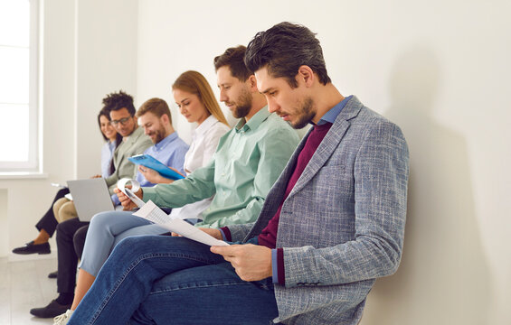 Group of young people sitting in line by a white office wall, waiting for a job interview or business meeting, reading, using laptops, and communicating. Business recruitment concept