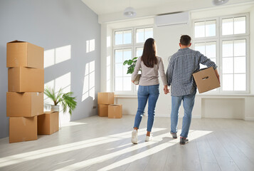 That's our new home. Family couple moving into modern house or apartment. Back view of young man and woman holding hands, carrying their things and walking into unfurnished living room with big window - obrazy, fototapety, plakaty