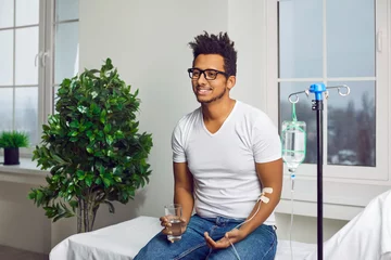 Fotobehang Happy, smiling young ethnic African American man sitting on a medical bed at the clinic, drinking water and receiving modern intravenous vitamin therapy through a sterile IV drip line infusion system © Studio Romantic