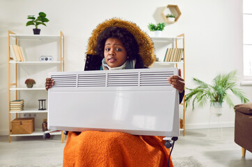African American young woman in winter jacket holding heater while sitting at home. Upset warmly...