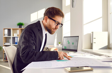 Professional surveyor in suit and glasses sitting at his table in office, working with modern city structure design plans, looking at cadastral maps, and studying plot numbers and boundaries