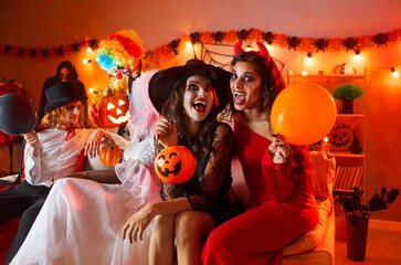 Portrait of two cheerful women in carnival costumes having fun during Halloween celebration at home. Women dressed as witch and devil sitting on sofa with balloon and pumpkin on background of people. - obrazy, fototapety, plakaty