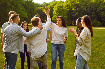 Team of happy, joyful diverse friends meet in the park on a good summer day. Happy man and woman give each other a high five while other young people are clapping hands. Teamwork concept