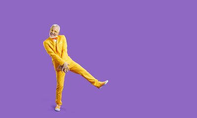 Funny banner ad. Cheerful eccentric and funny senior man in bright stylish clothes isolated on purple background. Full length of cool gray-haired man in yellow suit who is having fun near copy space.