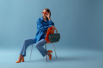 Fashionable young woman wearing trendy orange sunglasses, blue linen shirt, trousers, block heel shoes, holding straw wicker bag, posing on blue background. Studio portrait. Copy, empty space for text - 648665439