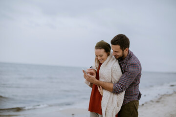 Young happy Caucasian couple using a smartphone while on on a sandy beach during winter