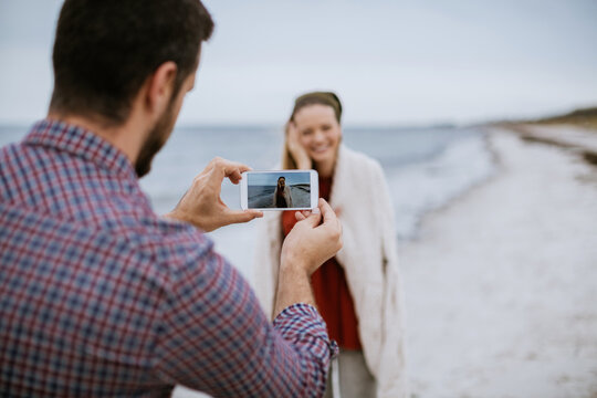 Young Caucasian man taking a picture of his wife on a smartphone while on a sandy beach during winter