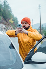 Young bearded man using phone while resting stop on road trip. He is wearing casual yellow jacket and red hat. Autumn travel by car.