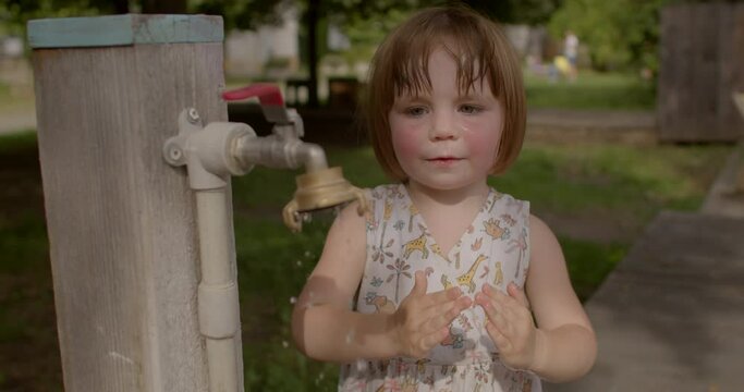 Toddler girl independently washing her face with water from an outdoor faucet during hot summer day. People lifestyle. Independence ability to perform basic self-care tasks without adult assistance.