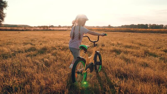 Little girl in helmet rides bicycle playing in sunset field country vacation