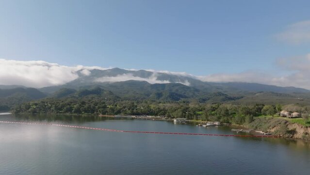 Aerial slide and pan of marina at bank of lake. Mountain ridge shrouded in clouds in background. Cachuma Lake, California, USA