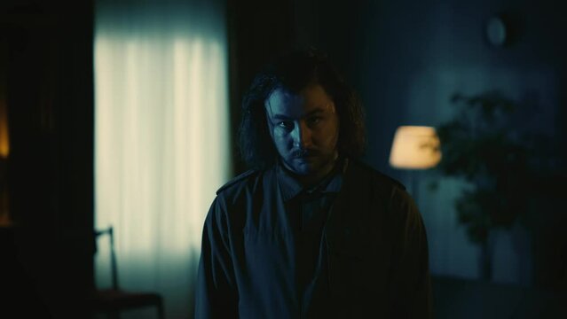 Man maniac with long hair standing in the dark room, looking directly at the camera with confident and straight face expression, tense atmosphere.