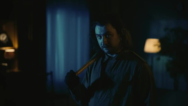 Man maniac with long hair standing in the dark living room, looking directly at the camera and holding baseball bat, thunderstorm outside.